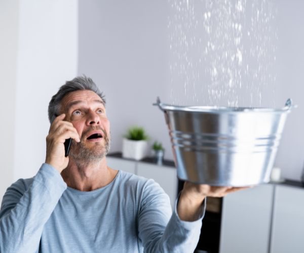 Person mit Eimer fängt Wasser auf und ruft Sanitär-Notdienst an